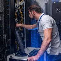 Server checking. Young bearded man in tshirt and jeans with badge in server room near open cabinet checking